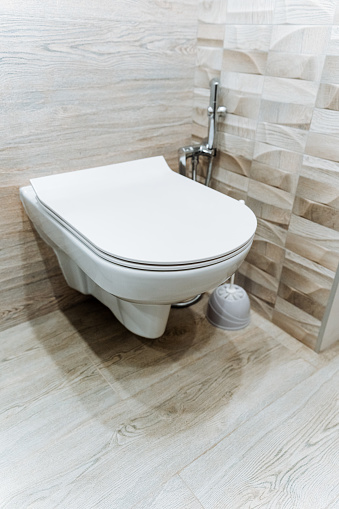 A white porcelain toilet with a rectangle shape is mounted to the wall in a bathroom. It is accompanied by a wood toilet seat and a bathroom sink. The floor is also mounted with plumbing fixtures