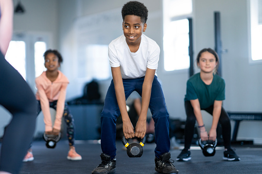 A small group of school age children are seen lifting kettlebells in a gym together.  They are each dressed comfortably in athletic wear and are focused on their squats as they participate in the class and follow the instructor.