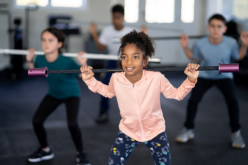 A small group of school aged children are seen lifting weight bars in a gym together.  They are each dressed comfortably in fitness attire and are focused on holding their front squat position while steadying the bar.