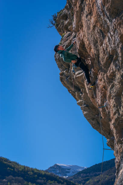 Caucasian man with black hair. Practicing rock climbing. Hanging from a single hand on an overhanging wall. With blue sky in the background