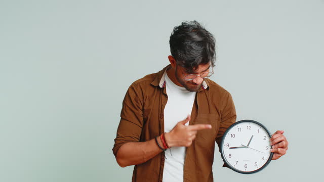 Indian young man showing time hour on wall office clock, ok, thumb up, approve, hurry up, deadline