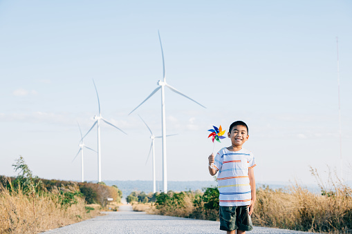 Cheerful child engaging with wind energy holding a pinwheel near turbines captures spirit of playful innovation. Showcases clean electricity amidst a countryside windmill farm under a serene blue sky.