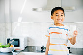 Smiling Asian little boy in kitchen holds milk cup. Portrait of cute son enjoying drink. Joyful child sips calcium-rich liquid, radiating happiness at home, Daily life health care Medicine food