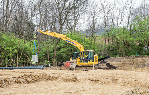 Dayton, Ohio - April 9, 2024: Construction worker uses backhoe to lower concrete block into trench of housing construction development drainage system.