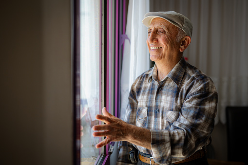 Careful elderly man standing by the window and removing the curtains in order to look at the view