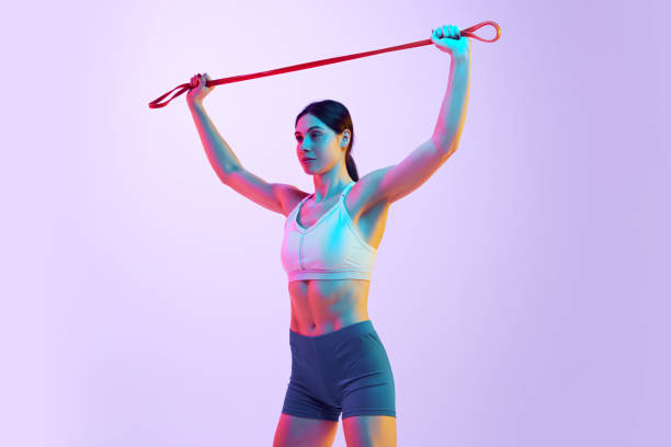 istock Young athlete woman in activewear doing exercises for hands using resistance band in neon light against gradient studio background. 2148842004