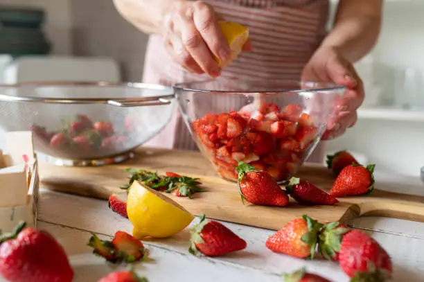 Woman marinating fresh chopped strawberries in a glass bowl. Healthy fruit dessert. Preparing food in the kitchen.