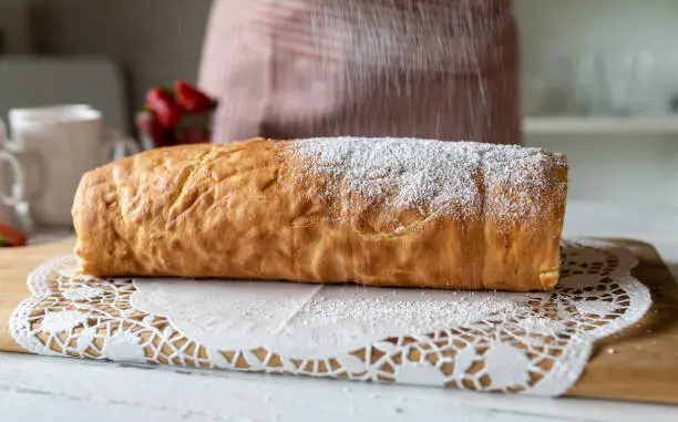 Sprinling powdered sugar over a fresh filled and baked cake roulade or swiss roll in the kitchen by a woman with apron