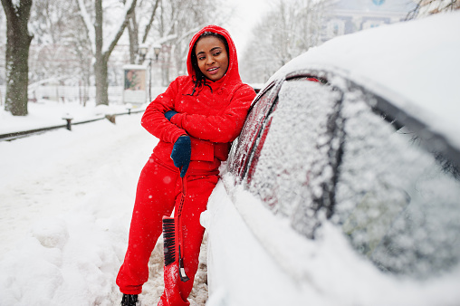 African american woman in red hoodie clean car from snow in winter day.