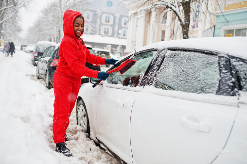 African american woman in red hoodie clean car from snow in winter day.
