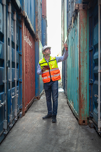 A logistics professional in safety gear is conducting an audit, verifying container numbers in a narrow corridor of a shipping yard.
