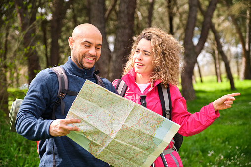 A man and woman are looking at a map together in a forest. The woman is pointing in a direction an the man is looking at the map.