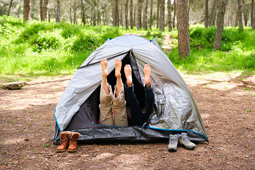 Two people are laying in a tent with their feet up. The tent is made of a grey material and is set up in a forest.