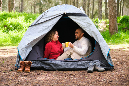 A couple is sitting in a tent, holding cups and enjoying each other's company. Scene is warm and intimate, as the couple shares a moment of relaxation and connection in the great outdoors.
