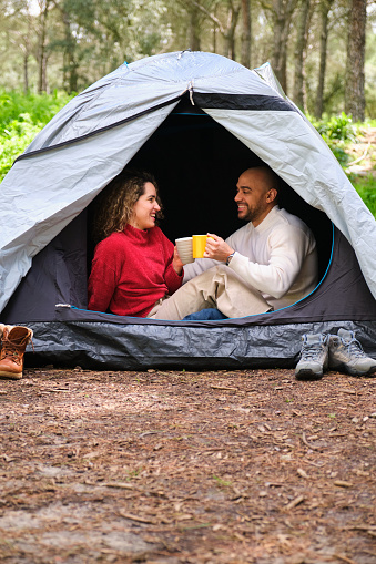 A couple is sitting in a tent, holding cups of coffee and smiling. Scene is warm and cozy, as the couple enjoys each other's company in a peaceful outdoor setting.