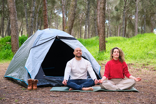 A man and woman are sitting by a tent in the woods meditating. They are both in a relaxed state, possibly meditating or enjoying the peaceful surroundings.
