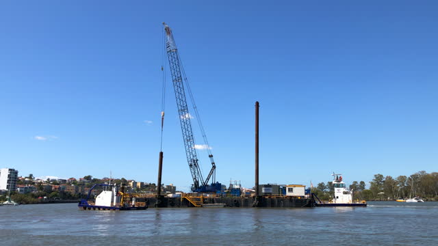 A Construction crane sitting on a barge being towed by two tug boats ready to build a bridge across a river