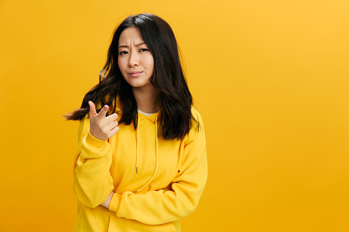 Hey, you. Confused incredulous cute Asian student young woman in yellow hoodie sweatshirt point finger at camera posing isolated on over orange studio background. Cool offer. People Emotions concept