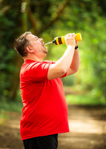 Close up image depicting a mid adult male jogger stopping to drink from his water bottle on a forest path. The man is so exhausted that he squirts water in his mouth and it soaks his face and shirt. The man is wearing sports training clothing - red t shirt and black shorts.