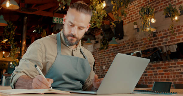 istock Owner, man and notes with laptop in restaurant for menu development or startup business planning for cafe. Person, online for research and marketing strategy for customers, profit sales or inventory. 2148848605