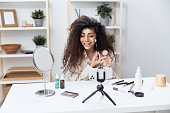 Smiling tanned handsome curly Latin beauty blogger in linen shirt how to put blush at makeup lesson in home interior. Copy space Mockup Banner. Influencer record video review blog using smartphone