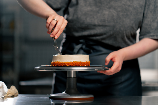 close up female baker in a professional kitchen distributes cream onto a sponge cake with a spatula