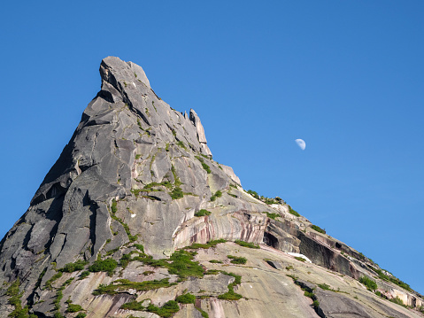 Moon over the sharp rock. Rock with a pointed top. Summer landscape with rock and blue sky.