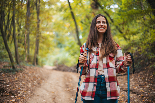 Beautiful woman traveler climbs uphill with a dog on a background of mountain views. She is with a backpack and in red clothes. Woman in the forest hiking with hiking poles