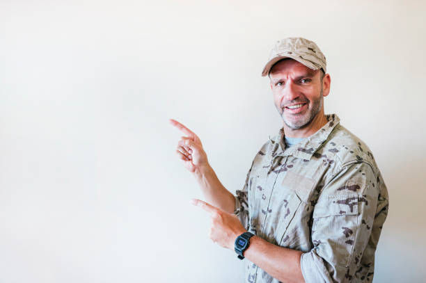 Handsome man in camouflaged army uniform smiling and looking at the camera pointing with two hands and fingers to the side.