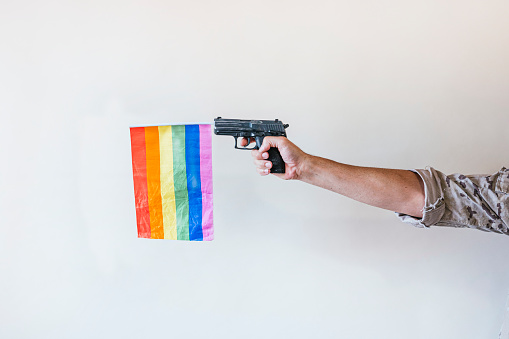 Hand of a military man carrying a gun with a multicolored pride flag.