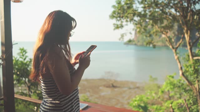 Young Woman Using Smartphone with Tropical Ocean View in Background.
