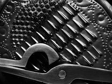 Black and white closeup of a stainless steel grater and a pair of stainless steel tongs the most useful items in the Indian (South Asian) kitchen.