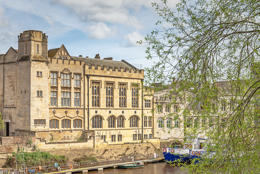 York, UK. April 14, 2024.  The 15th Century Guildhall in York. The historic building stands beside a river and two tour boats are moored alongside.  A sky with clouds is above and a tree is in the foreground.