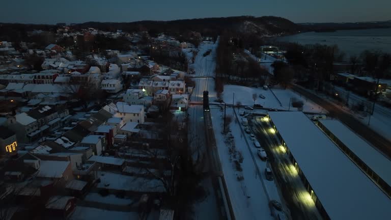 Aerial birds eye shot of train on snowy track in american town at night. Lighting buildings and lantern in winter scene. Suburb district of small town in USA. Drone flyover.