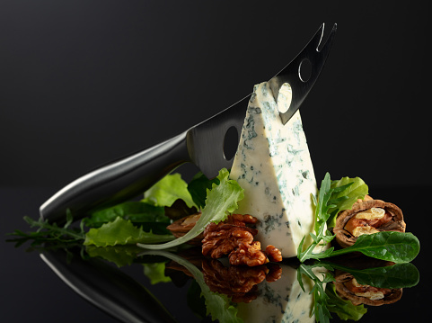 Blue cheese with knife, walnuts and fresh greens on a black background. Copy space.