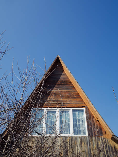 An attic window offers a glimpse into  cozy interior. In the foreground, bare tree branches frame the scene, their delicate silhouettes adding a touch of natural elegance. Behind the house, the clear blue sky serves as a serene backdrop, while white lace pattern curtains softly billow from the attic window, exuding a sense of rural charm in the fresh embrace of early spring.