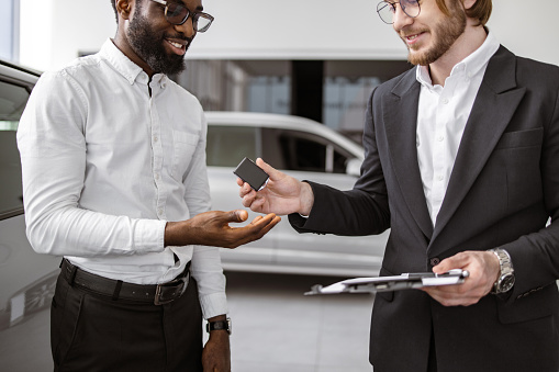 Cropped view of Caucasian sales manager in suit giving car key to young Caucasian businessman standing beside new modern vehicle. Choosing and buying new car concept.