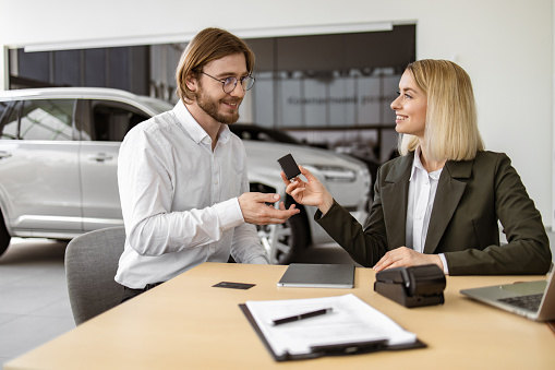 Happy Caucasian young man sitting at table and receives keys of SUV car from confident blond saleswoman in suit in bright showroom. Concept of buying new modern vehicle.