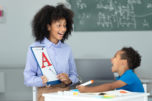 Cheerful black female speech therapist and little boy learning and pronouncing letters during lesson in office