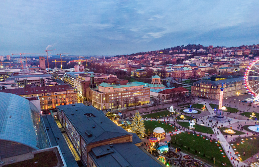 Drone point of view on Shlossplatz of Stuttgart - soon Christmas or maybe another holiday. People eating ,
 drinking and enjoying life. Illuminated city in twilights . In left upper corner - several cranes- machinery  maybe there Main railroad 
station of Stuttgart