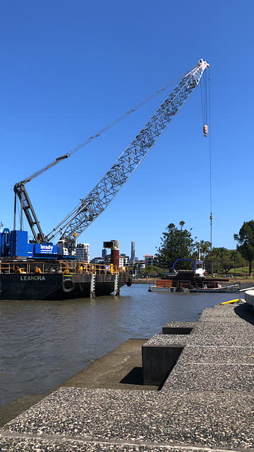 Construction crane on a large barge slowly lowering its boom to a tugboat on the river below at a bridge construction site