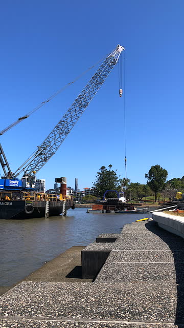 Construction crane on a large barge slowly lowering its boom to a tugboat on the river below at a bridge construction site