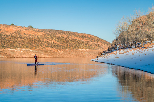 male paddler in red drysuit is paddling a stand up paddleboard on mountain lake in Colorado, winter scenery