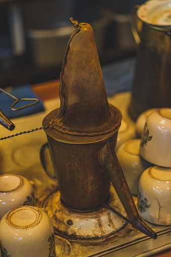 A gleaming traditional copper coffee pot sits on a stovetop next to a stack of ceramic mugs. The pot has a classic design with a rounded body and a long spout. Steam is gently rising from the pot, indicating that fresh coffee is brewing.