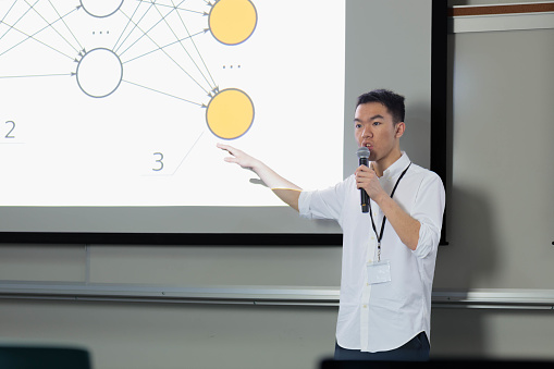 Teacher teaching about artificial intelligence in college, gesturing at the screen with his hands while holding a microphone, with a neural network infographic displayed on a large projection screen