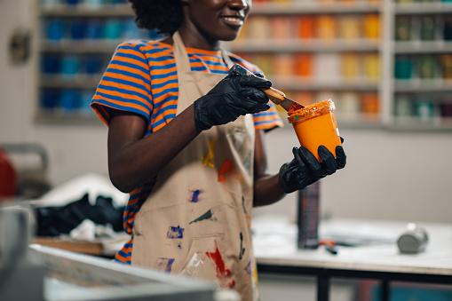 Cropped picture of multicultural female graphic technologist with bucket of orange paint, mixing paint and preparing it for printing.Close up of african american industrial worker's hands mixing color