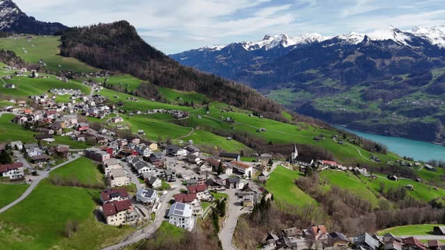 Drone shot showing idyllic village on green mountain and snowy alps with lake in background. Aerial orbiting wide shot.