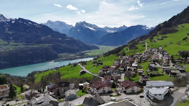 Quaint Swiss Landscape with buildings and homes on hill. View on turquoise Lake Walen and snowy summits of mountains. Aerial panning wide shot.