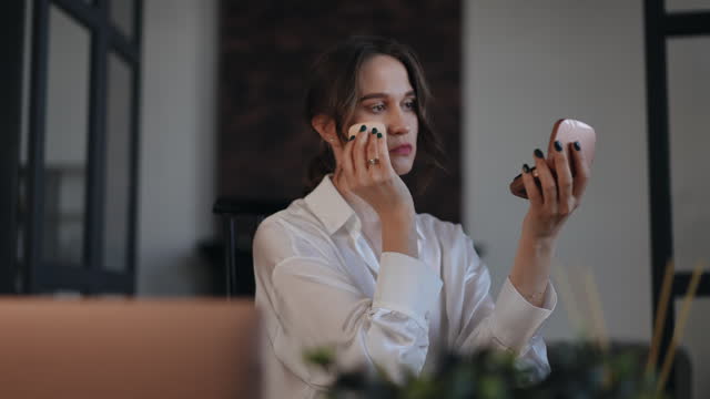 Business woman doing makeup while sitting on a chair in her office. A woman looks at herself in the small mirror of a powder compact and powders her face.
