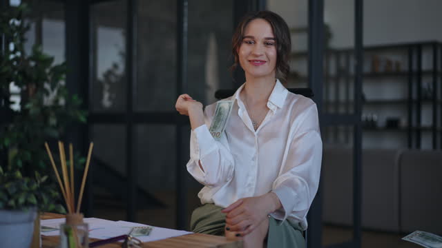 Happy smiling businesswoman looking at camera and waving fan of American dollars in hand while sitting at desk in office. The woman thought and throw up the money she was holding in her hand.